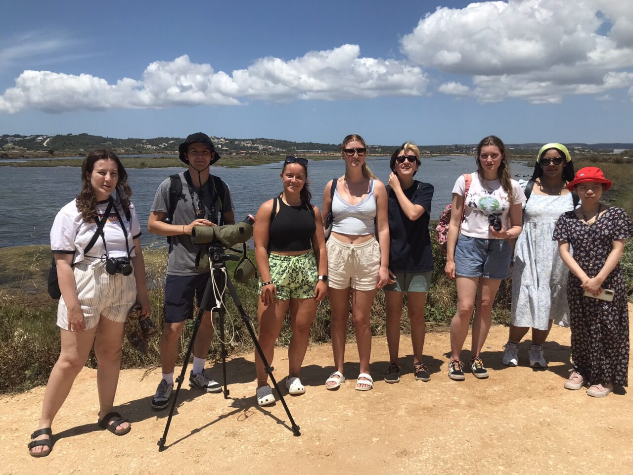 Grŵp o bobl (â chamera ar drybedd) sy’n sefyll mewn rhes. Mae dŵr yn y cefndir ac awyr las ynghyd â rhywfaint o gymylau.