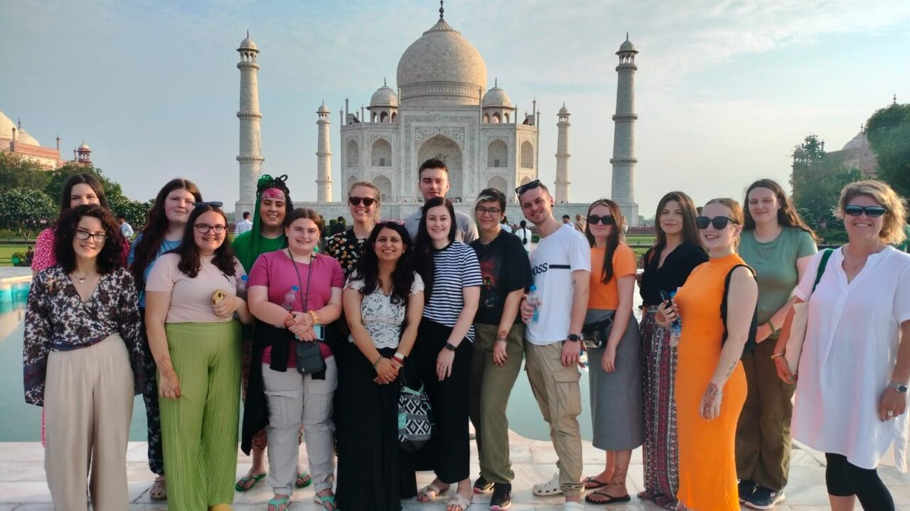 Criw o bobl yn sefyll o flaen y Taj Mahal ar ddiwrnod heulog. A group of people stood in front of the Taj Mahal on a sunny day.