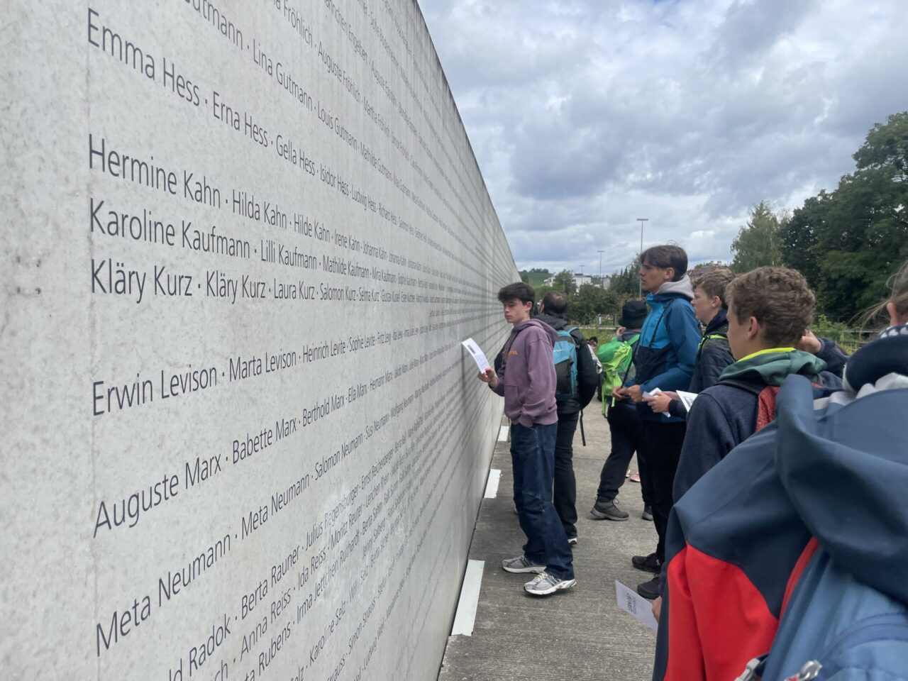 Grŵp o bobl ifanc yn eu harddegau yn sefyll o flaen cofeb o’r ail ryfel byd. Mae'r gofeb yn llawn enwau ac mae'r arddegau i'w gweld yn darllen y rhain.