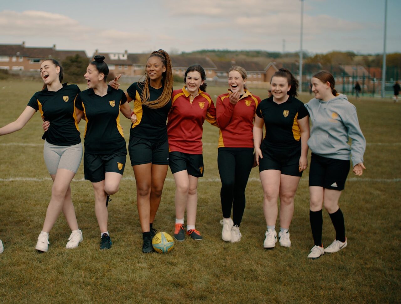 Saith o bobl yn sefyll mewn llinell yn gwenu ac yn chwerthin. Maen nhw'n gwisgo gwisg rygbi ac mae 'na bêl rygbi ar y llawr.