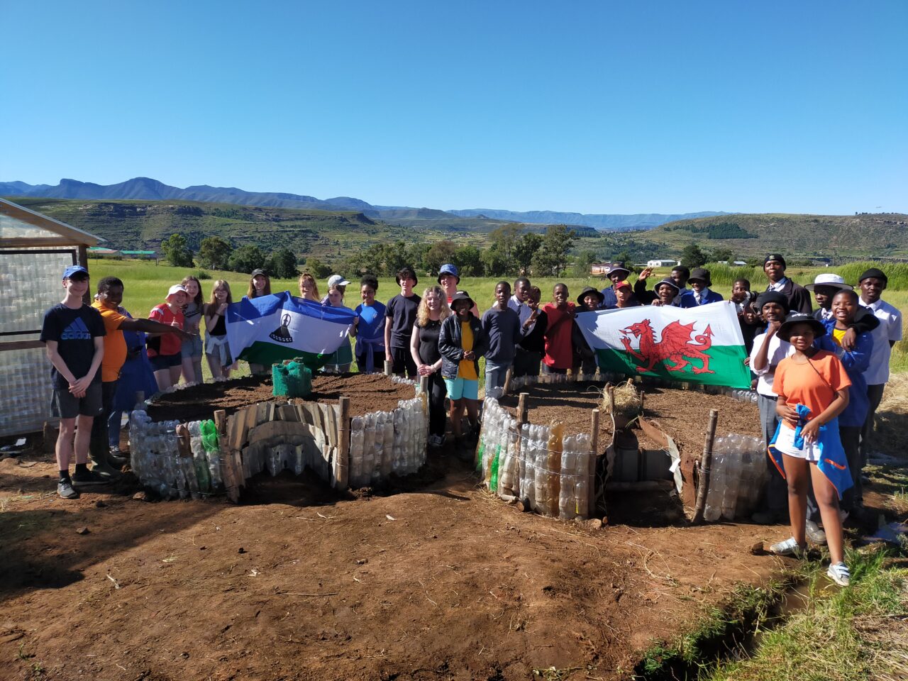 A large group posing for a photo and holding Welsh and Lesotho flags. There is a clear blue sky and green spaces in the background.