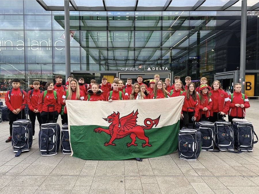 criw mawr o bobl ifanc yn sefyll y tu allan i faes awyr gyda'u bagiau yn dal baner fawr Cymru / a large group of young people standing outside an airport with their luggage holding a large Welsh flag