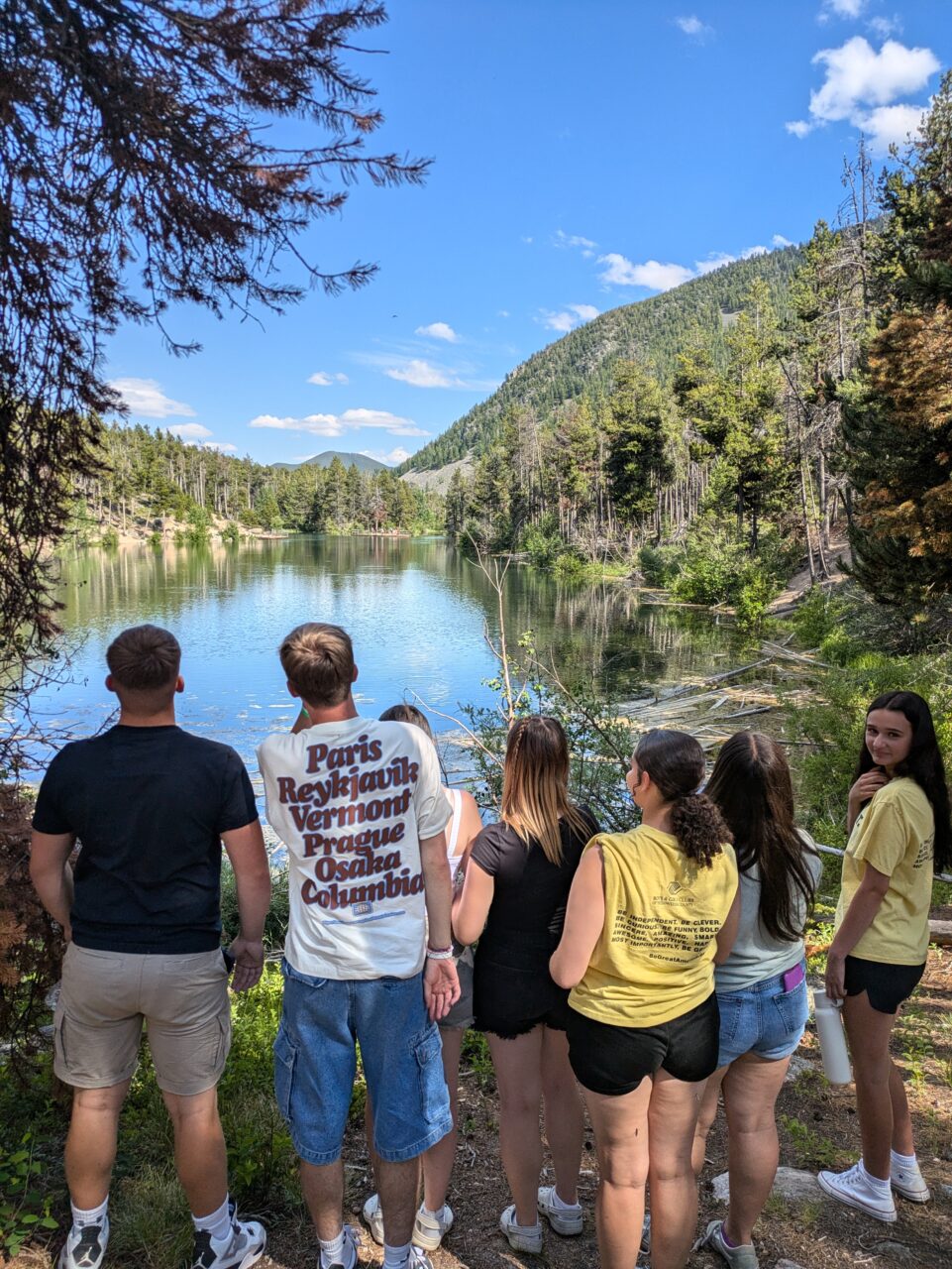 Saith o bobl yn edrych allan dros lyn mawr wedi'i amgylchynu gan goed ar ddiwrnod heulog. Mae mwyafrif y grŵp yn wynebu'r llyn, ond mae un wedi troi at y camera ar gyfer y llun / Seven young people looking out over a large lake surrounded by trees on a sunny day. Most of the group are facing the lake, but one has turned to face the camera for the photo