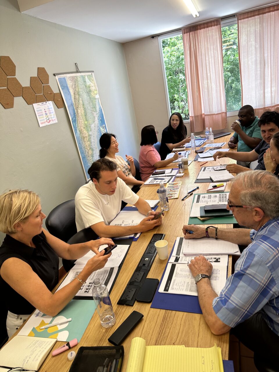 Naw oedolyn yn eistedd o amgylch bwrdd hir gyda llyfrau gwaith o'u blaenau. Maent yn cael sgyrsiau mewn grwpiau bach / Nine adults sat around a long table with workbooks in front of them. They are all having small group conversations