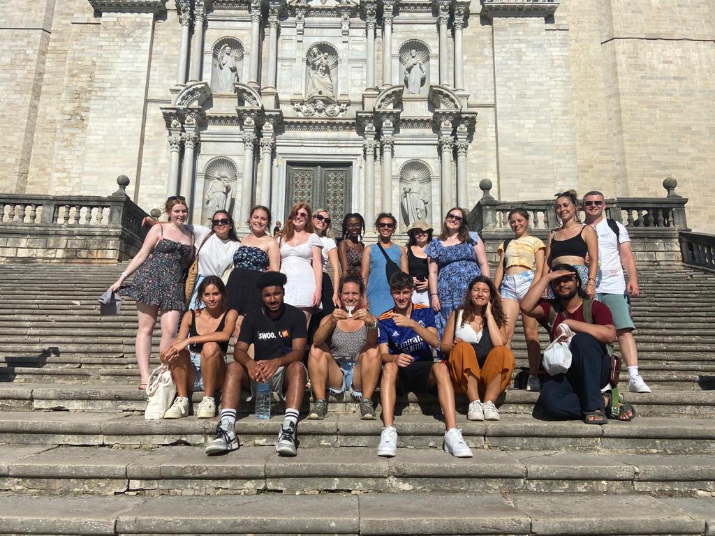 Grŵp o tuag at 20 oedolion ifanc o flaen addoldy. Mae'r bobl yn y rhes flaen yn eistedd ar stepiau yr addoldy ac y mae'r gweddill yn sefyll y tu ôl iddyn nhw. / A group of around 20 young adults in front a a cathedral. The front row of people are sat on the steps in front of the cathedral, and the rest are stood behind them.