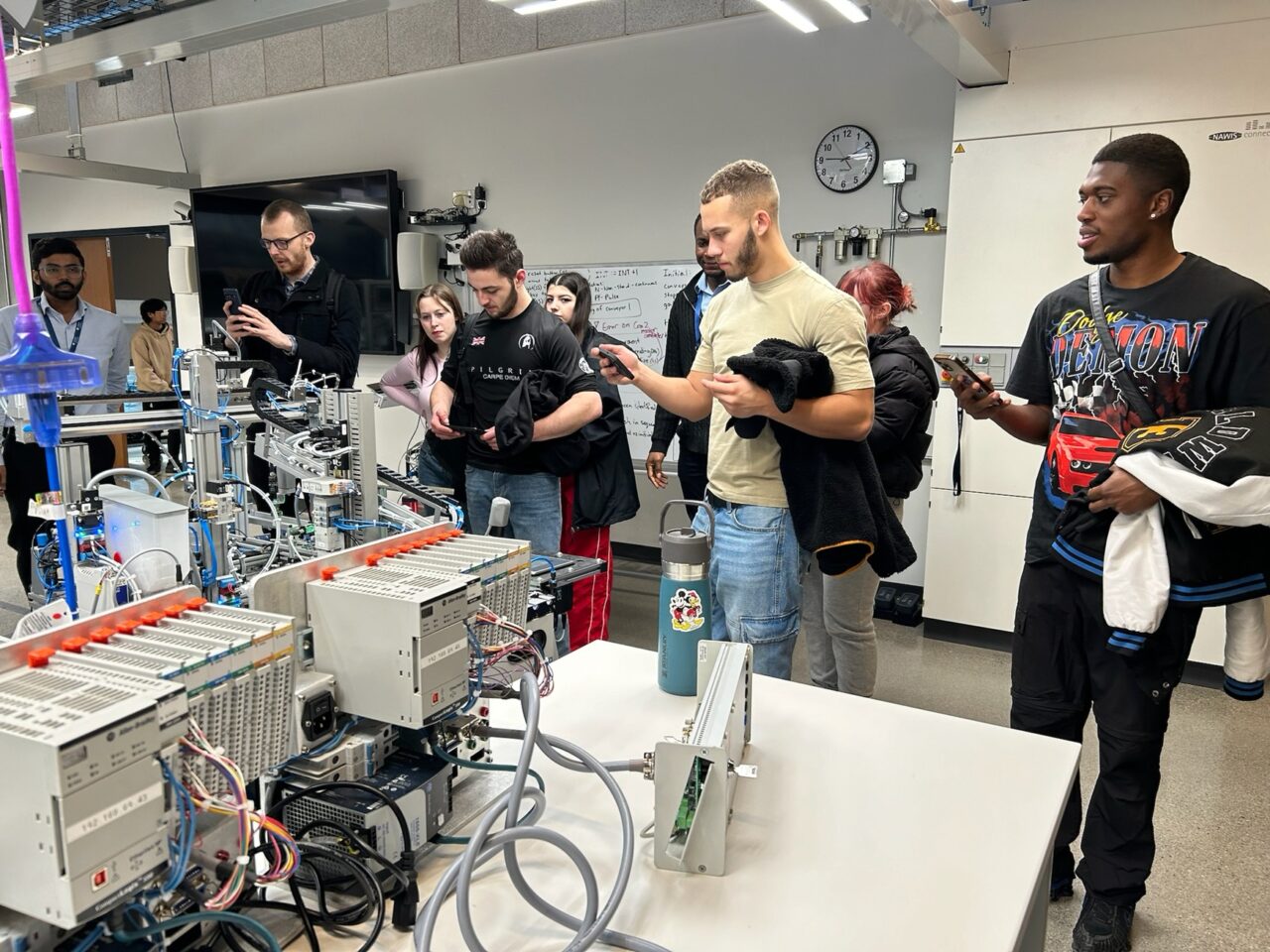 grŵp o fyfyrwyr yn edrych ar dechnoleg mewn ystafell fawr / a group of students looking at technology in a large room