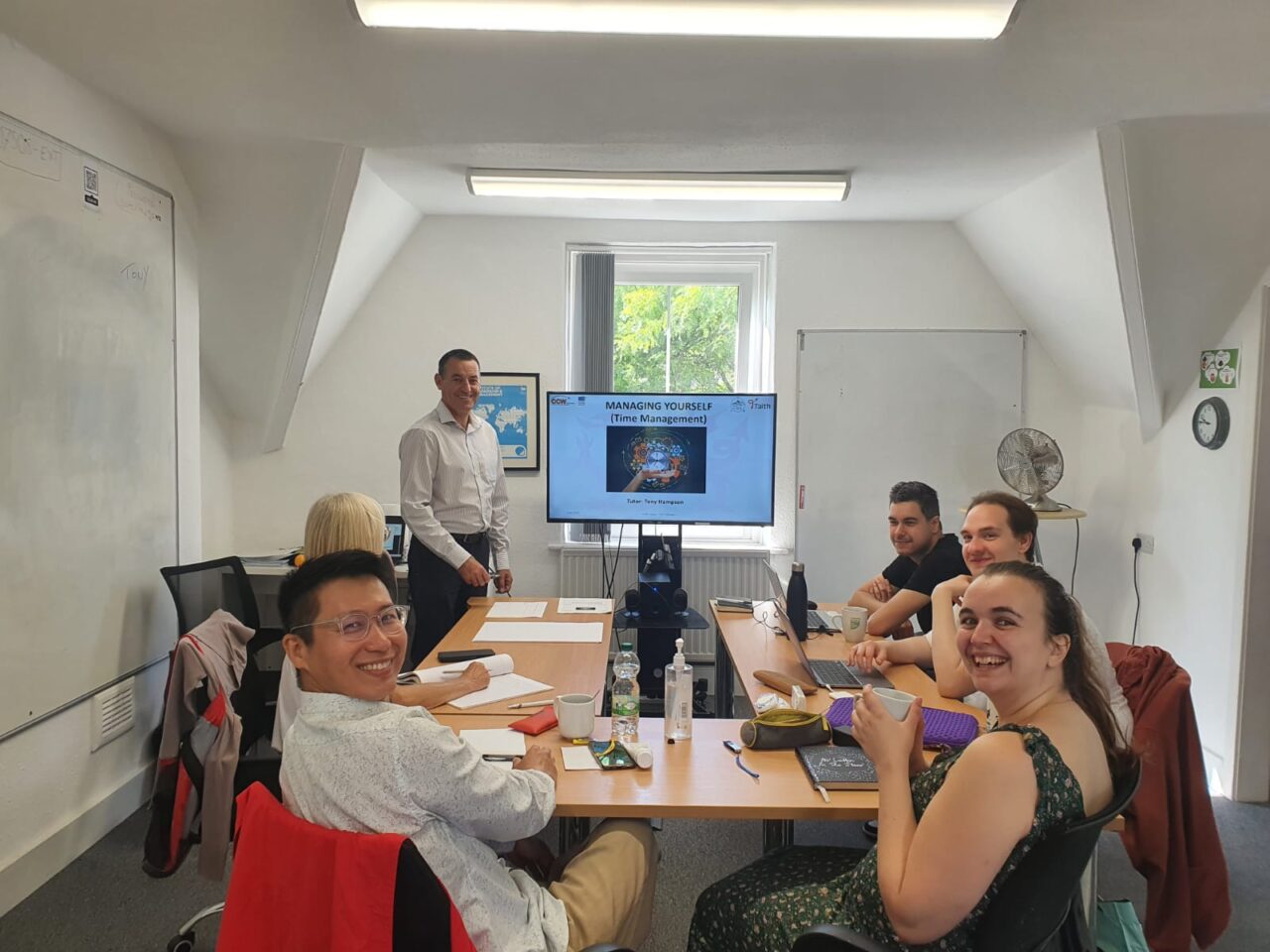 Grŵp o chwe oedolyn yn eistedd mewn ystafell ddosbarth fechan o amgylch byrddau gyda chyflwyniad o'u blaenau / A group of six adults in a small classroom sat around tables with a presentation in front of them.