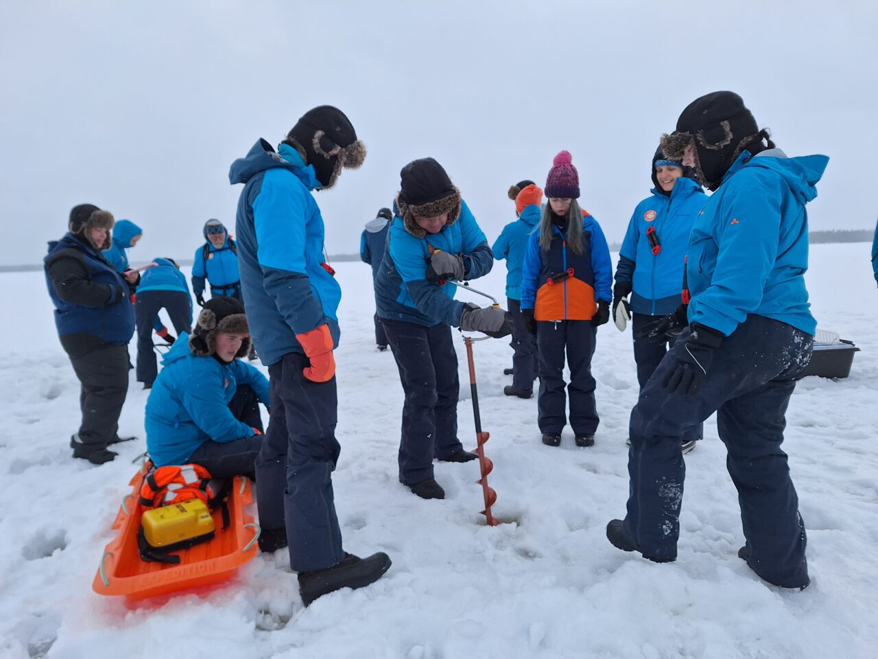 Criw o bobl ifanc ac oedolion yn sefyll ar iâ yn edrych ar un person yn drilio twll drwy'r rhew / A group of young people and adults standing on ice looking at one person drilling a hole through the ice
