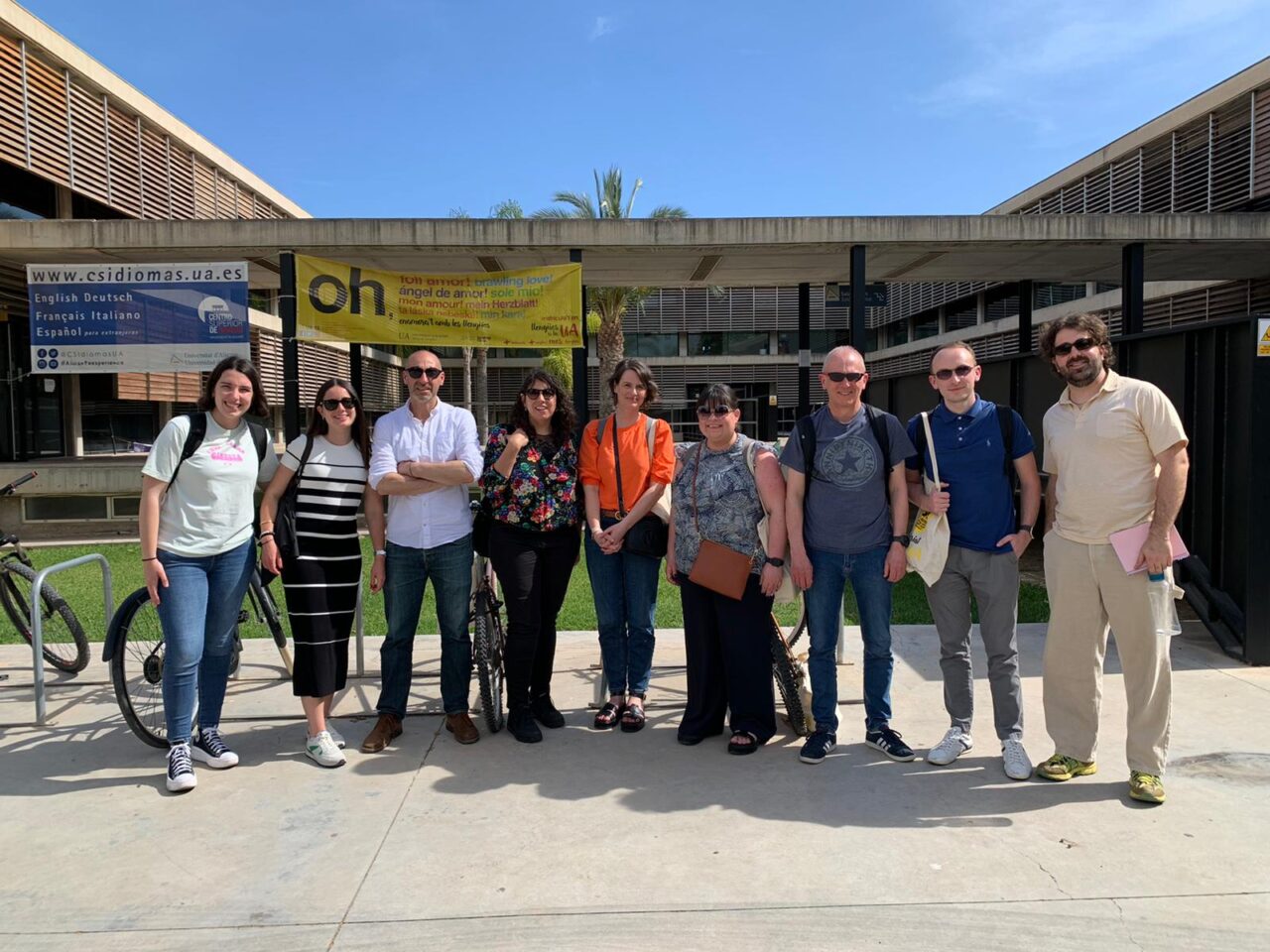 Naw oedolyn mewn llinell yn sefyll am lun y tu allan i adeilad. Mae awyr las yn y cefndir / Nine adults standing in a line posing for a photo outside a building. There is a blue sky in the background