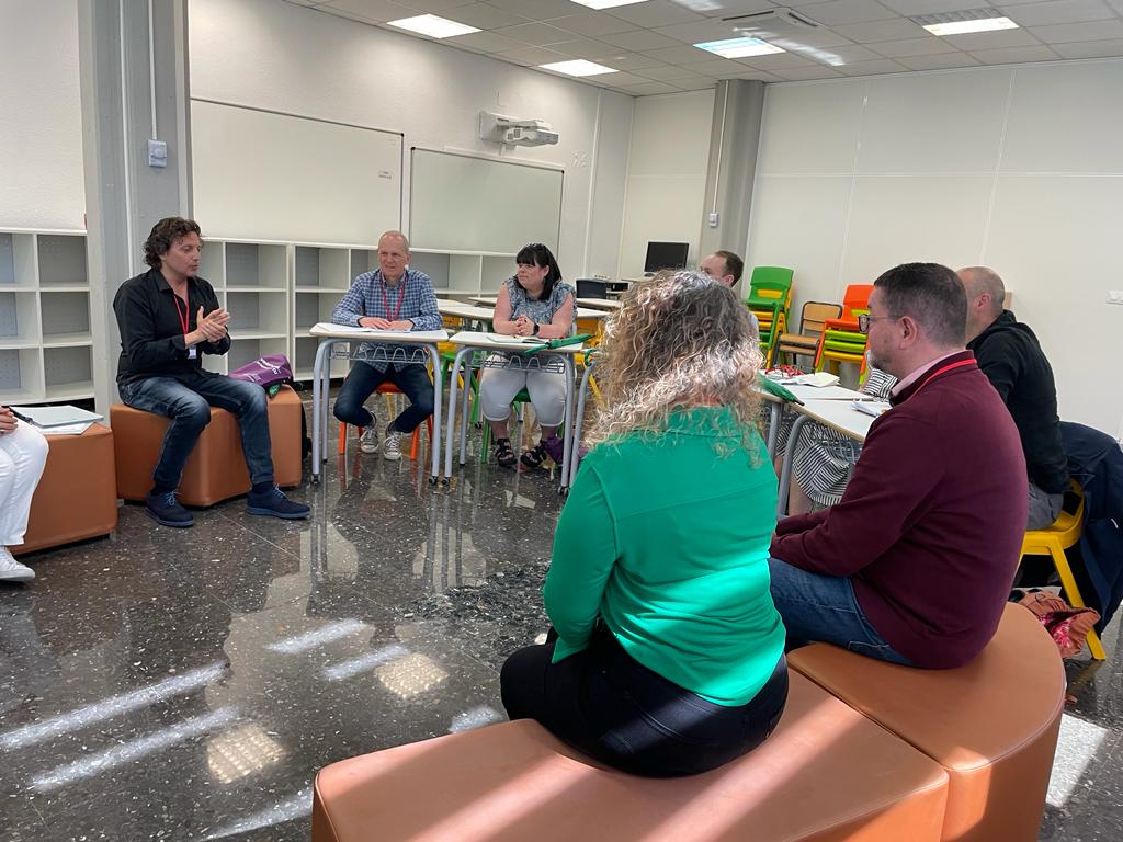 Saith o bobl yn eistedd mewn ystafell ddosbarth yn cael trafodaeth. / Seven people sat down in a classroom setting having a discussion.