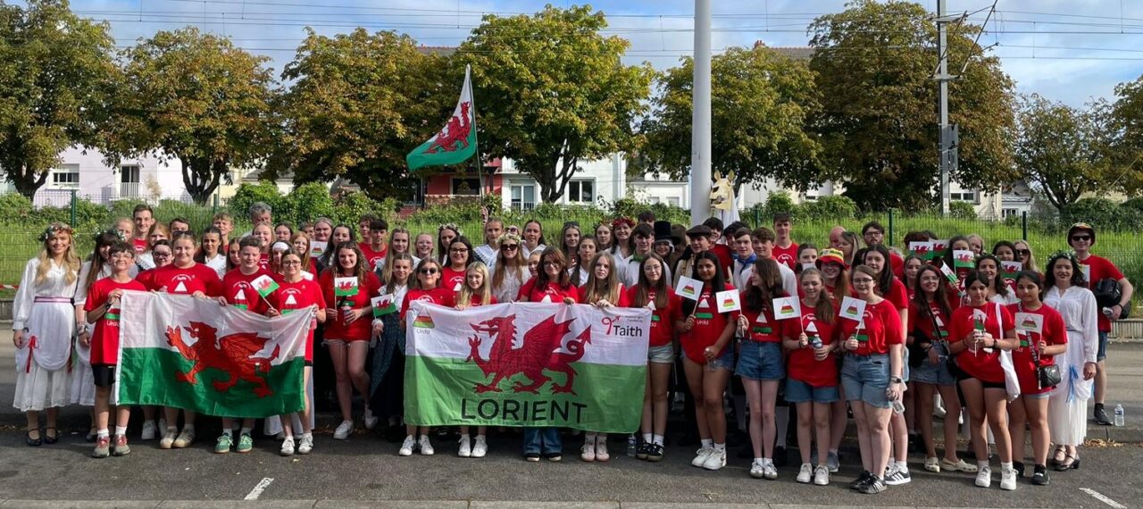 Grŵp mawr o ferched a bechgyn mewn crysau-T coch yn gwenu at y camera. Mae rhai o'r merched yn gwisgo gwisg werin wyn a chylch o flodau yn ei gwallt. Mae'r rhan fwyaf ohonyn nhw'n chwifio fflagiau bach baner Cymru neu logo'r Urdd. Mae'r merched yn y blaen yn dal baner mawr Cymru gyda 'Lorient' o dan y ddraig gyda'r logo Urdd ar yr ochr chwith a'r logo Taith ar yr ochr dde.