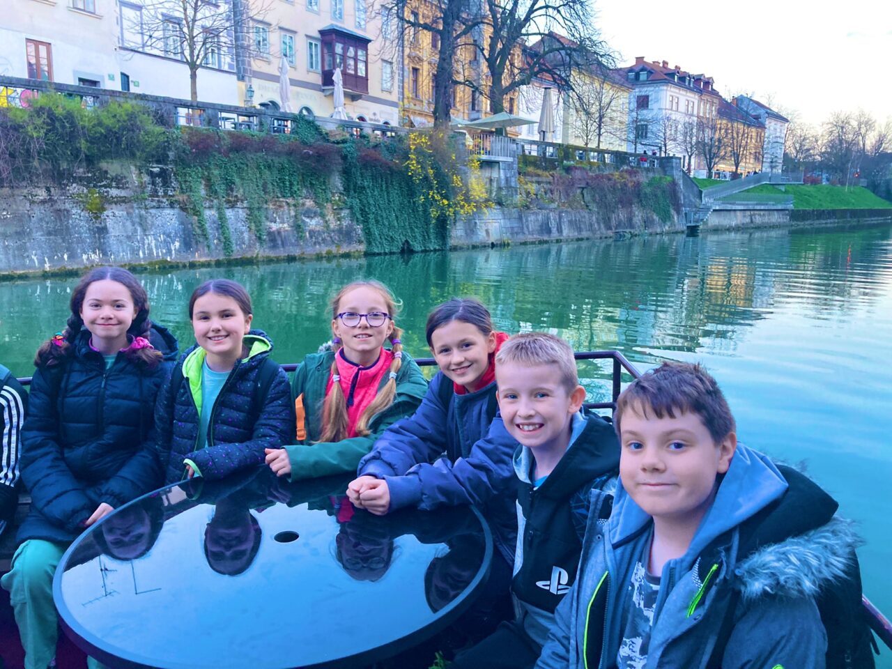Grŵp o blant ysgol gynradd yn gwenu at y camera wrth eistedd o gwmpas bwrdd ar bad ar afon. Y mae yna nifer o dai lliwgar wrth lan yr afon. A group of primary school children sat around a table on a boat on a river and smiling at the camera. There are colourful buildings along the banks of the river.