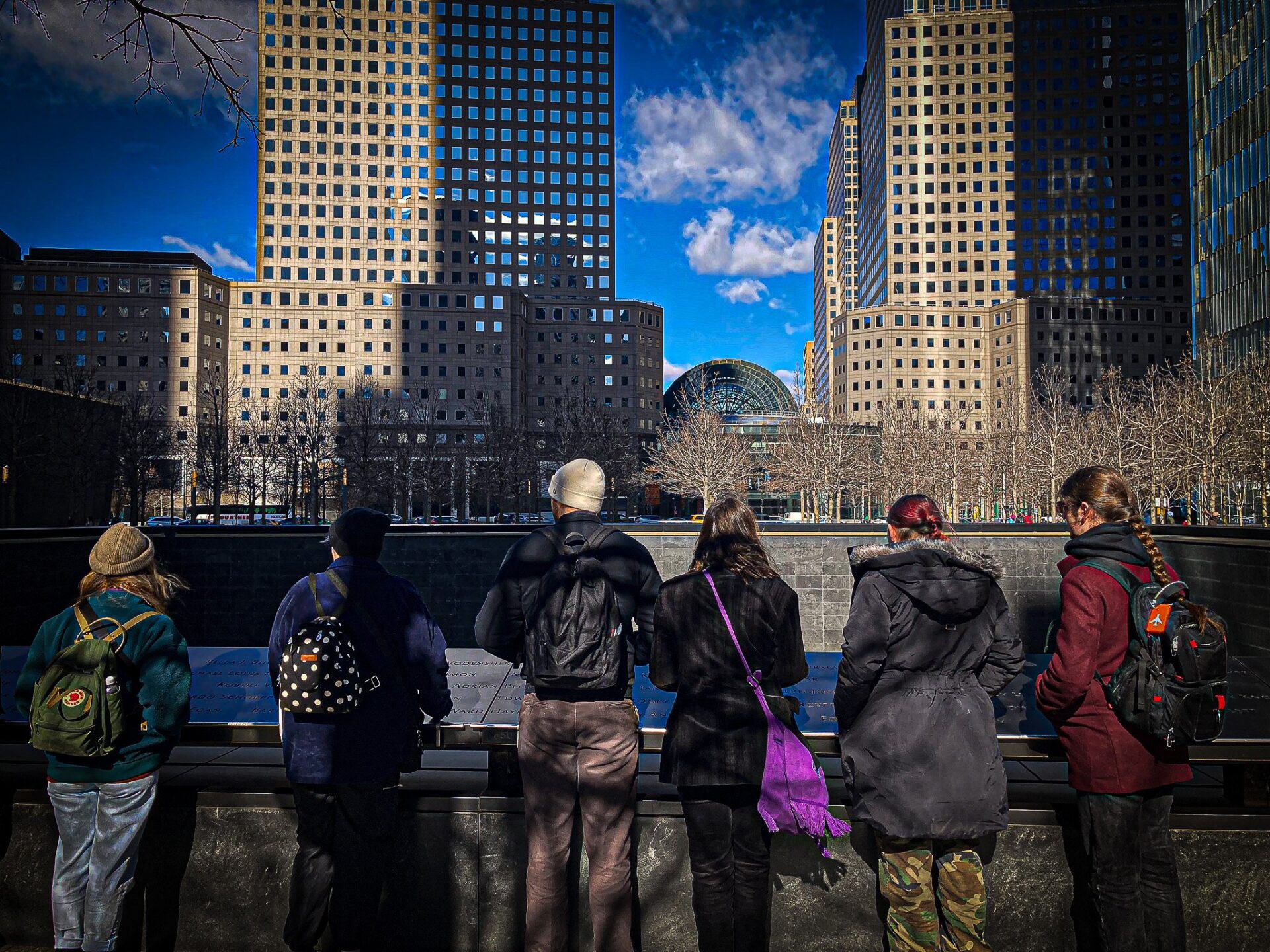 Chwech o fyfyrwyr a’u cefnai i’r camera yn edrych ar gofeb Medi 11eg yn Efrog Newydd. Y mae yna nifer o entrychdai wedi e’u hadlewyrchu yn erbyn yr awyr las dwfn yn y cefndir. Six students facing away from the camera looking at the September 11 memorial in New York. There are a number of skyscrapers reflected against a deep blue sky in the background.