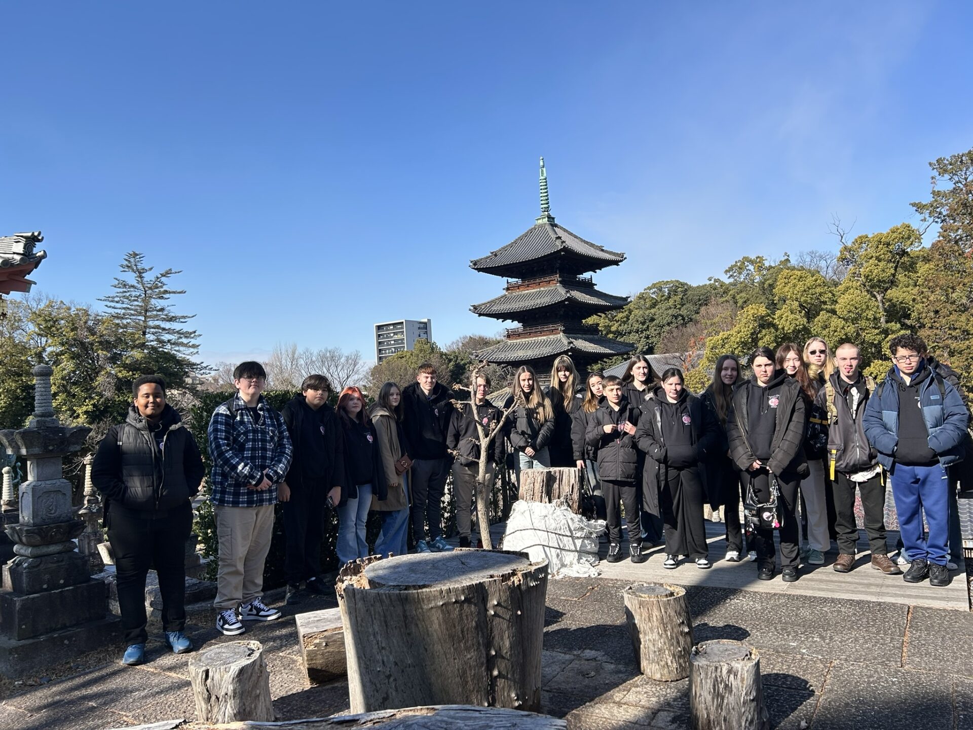 Grŵp mawr o ddisgyblion yn sefyll y tu allan, o bosibl mewn parc Siapaneaidd yn sefyll am lun. O'u blaenau mae ardal eistedd gron wedi'i gwneud o foncyffion coed a'u hôl mae adeilad bach Siapaneaidd.
