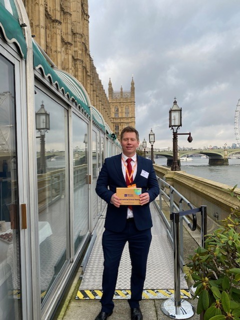Athro Andy Wallis yn sefyll y tu allan i Dŷ'r Senedd yn dal lefel Achrediad Gwobr Ysgolion Ryngwladol y British Council