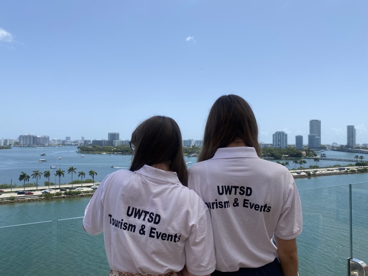 Dau fyfyriwr yn edrych dros falconi ar olygfa o ddŵr ac adeilad yn y cefndir. Dim ond cefn y myfyrwyr allwch chi eu gweld ac mae 'UWTSD Tourism & Events' wedi'i argraffu ar eu crysau.