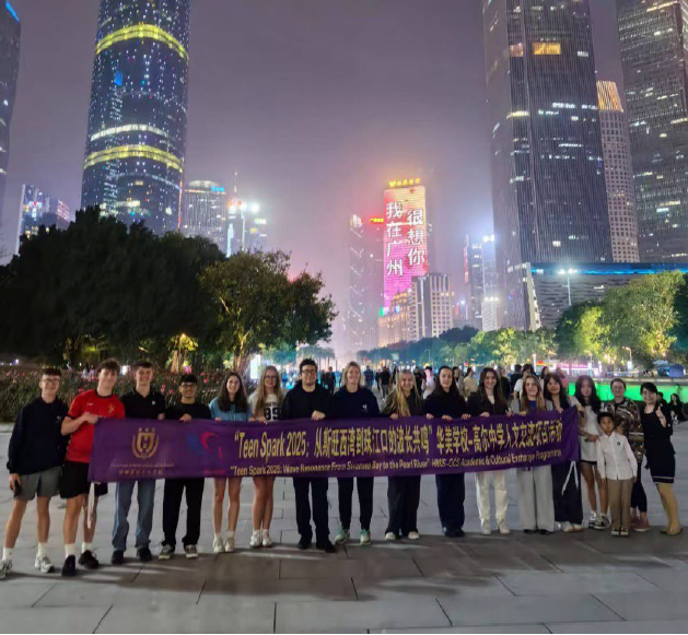 Cyfranogwyr Teen Spark 2025 yn dal baner y rhaglen mewn sgwar dinas yn y nos, gyda nendyrau wedi'u goleuo a chefndir gorwel yn Guangzhou.