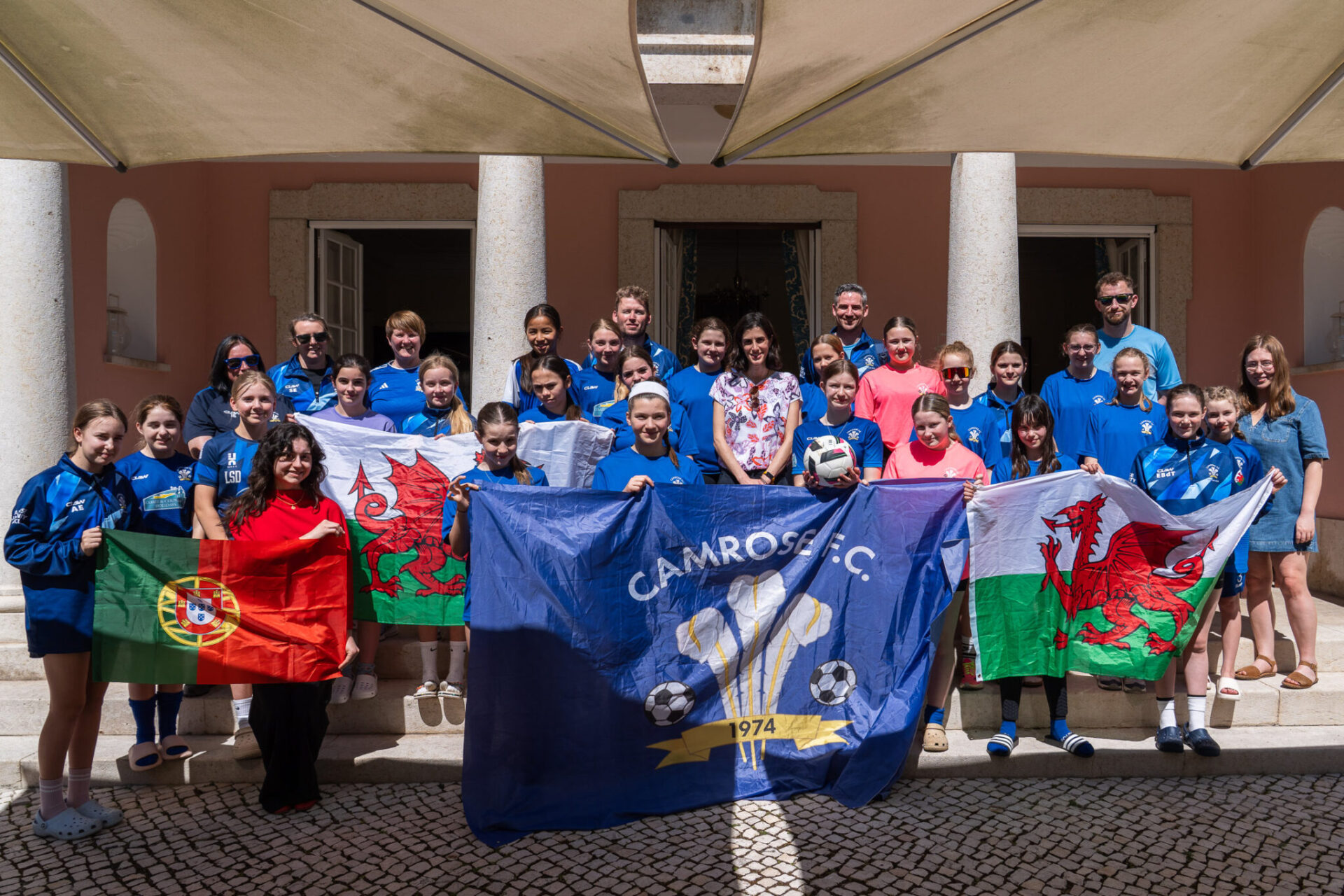 Mae grŵp o chwaraewyr pêl-droed ifanc a hyfforddwyr o Glwb Pêl-droed Camrose yn sefyll tu allan i adeilad, yn dal baneri Cymru, Portiwgal a chlwb yn ystod ymweliad cyfnewid chwaraeon rhyngwladol.