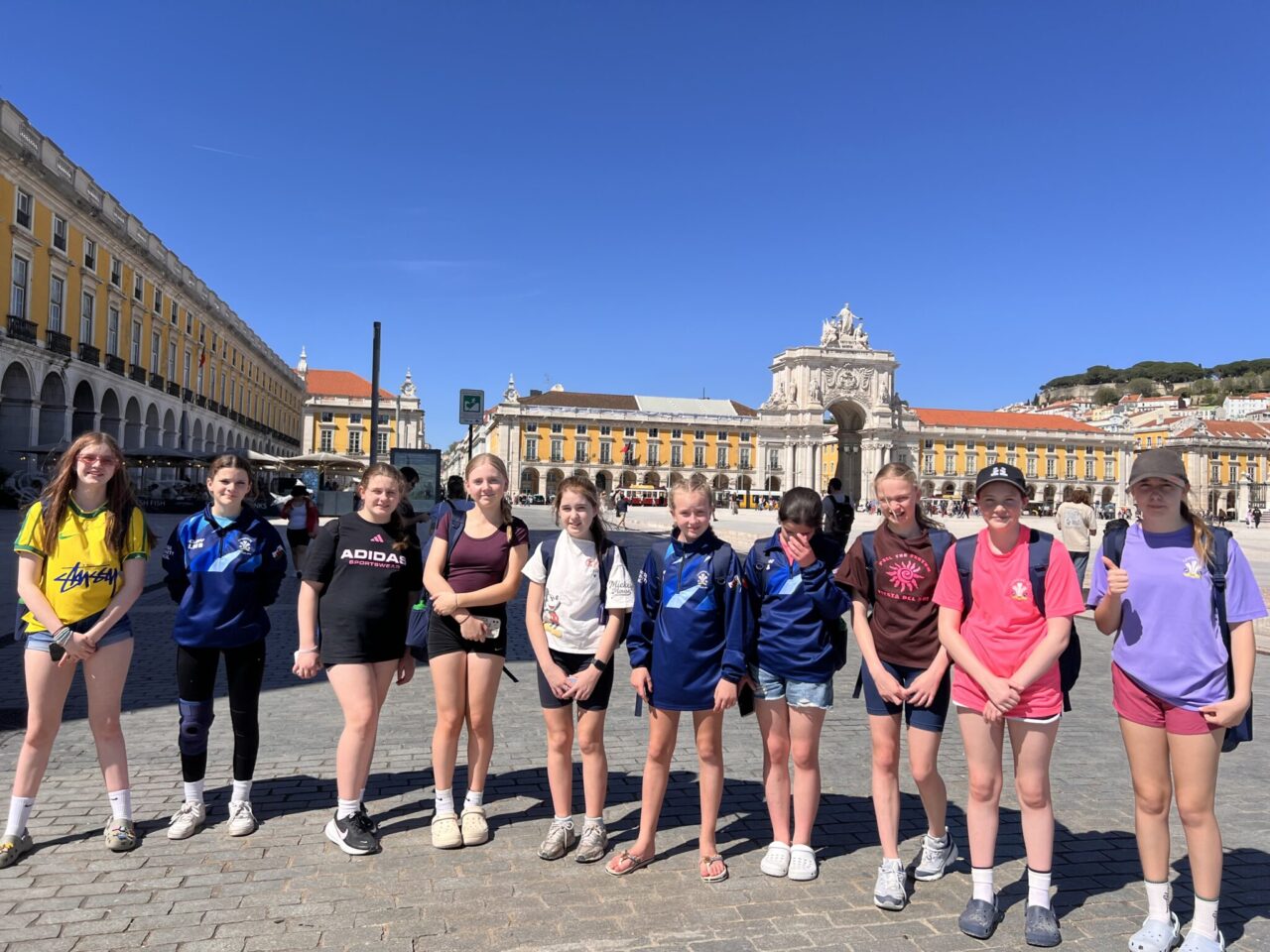Cyfranogwyr ifanc o Glwb Pêl-droed Camrose ar ymweliad cyfnewid rhyngwladol a gefnogir gan Taith yn sefyll yn Praça do Comércio, Lisbon, gyda bwa Arco da Rua Augusta a'r adeiladau hanesyddol cyfagos tu ôl iddyn nhw.