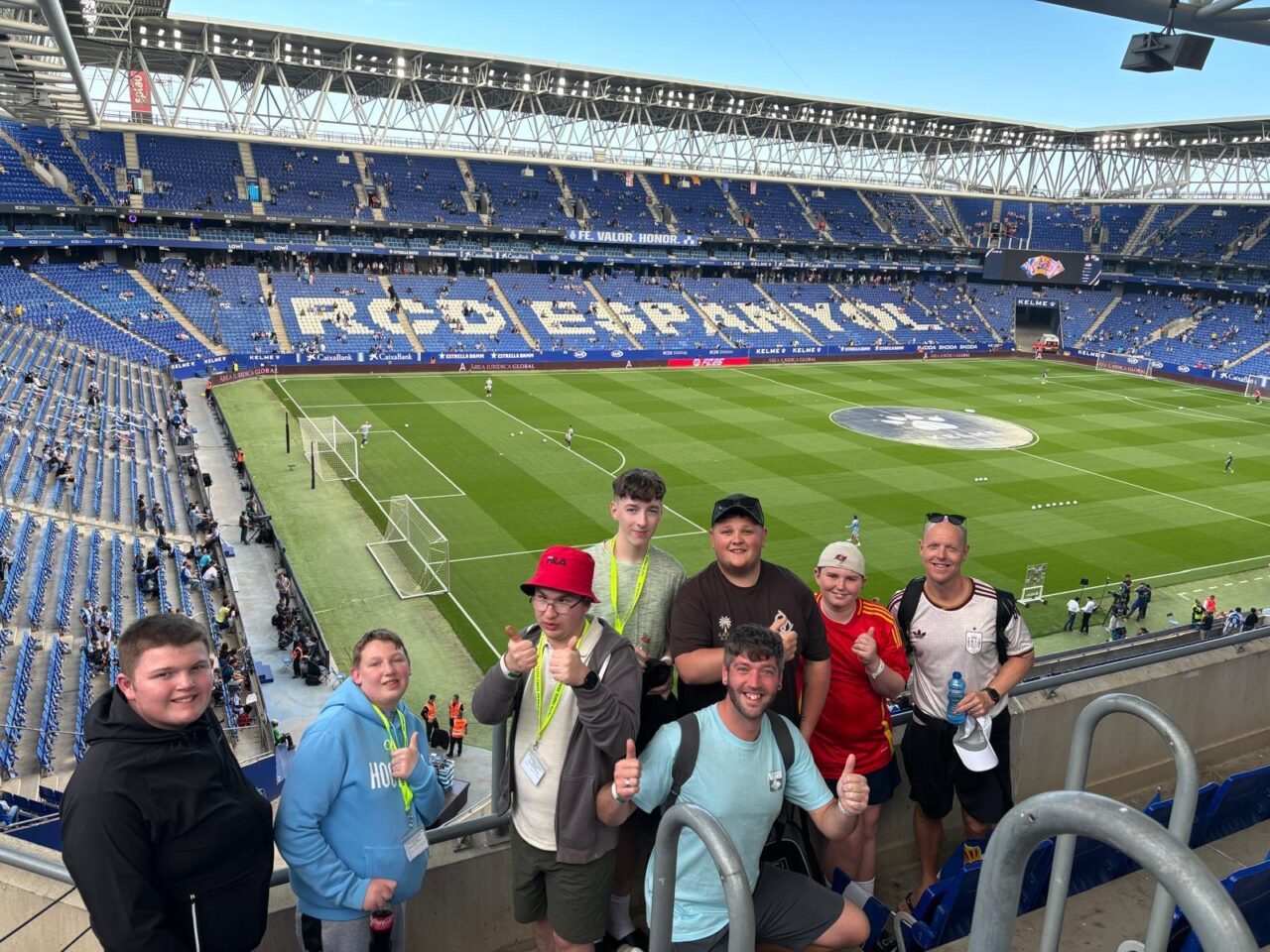 Grŵp bach yn sefyll yn eisteddleoedd stadiwm pêl-droed RCD Espanyol, gyda'r cae a'r seddi glas i'w gweld yn y cefndir.