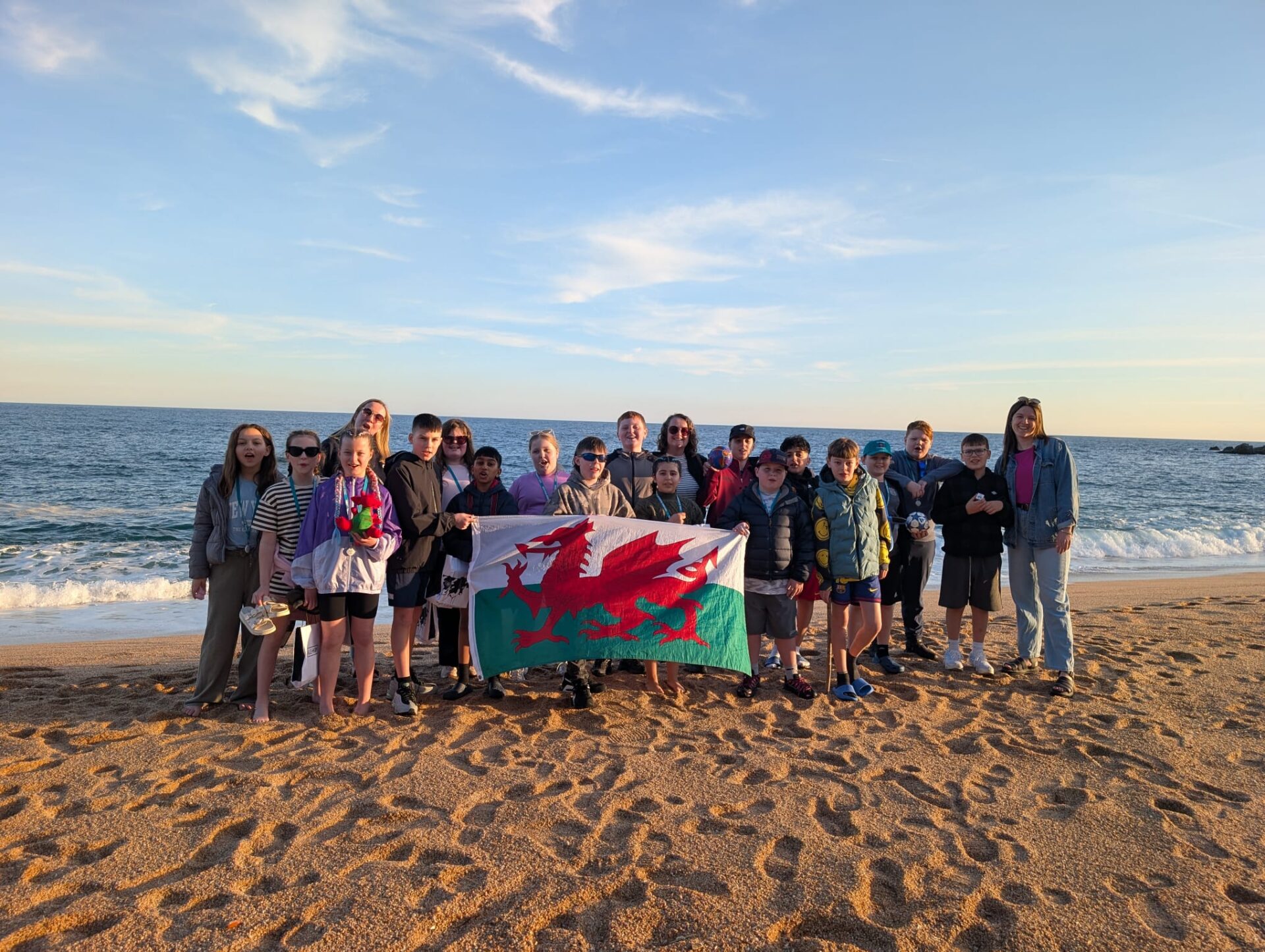Grŵp o ddisgyblion Blwyddyn 6 o Ysgol Alexandra ar draeth yn Barcelona yn dal baner Cymru, yn cymryd rhan mewn prosiect symudedd rhyngwladol a ariennir gan Taith.