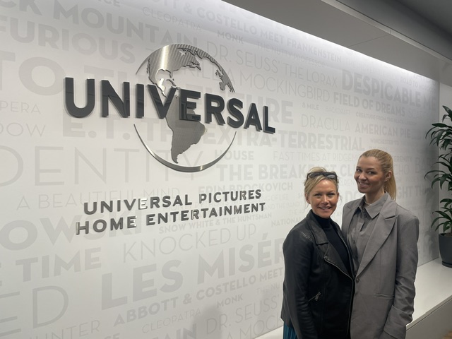 Marlen Komorowski a Sara Pepper yn sefyll o flaen wal yn arddangos logo Universal Pictures Home Entertainment tu mewn i adeilad swyddfeydd.